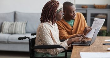 Side view portrait of young African-American woman using wheelchair working from home with husband helping her copy space